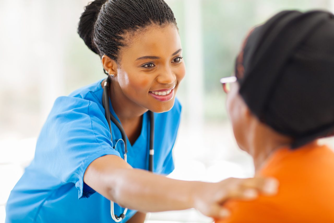 A nurse in scrubs leaning in and smiling as she reassures an older patient